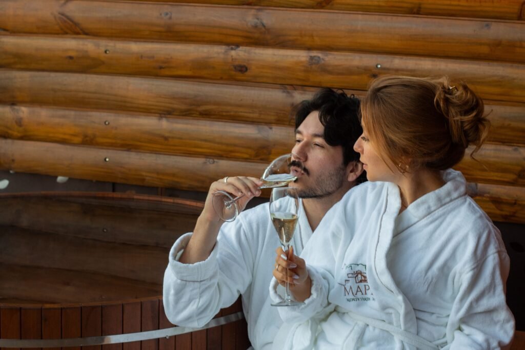 A relaxed couple in robes sipping champagne inside a rustic wooden sauna setting.
